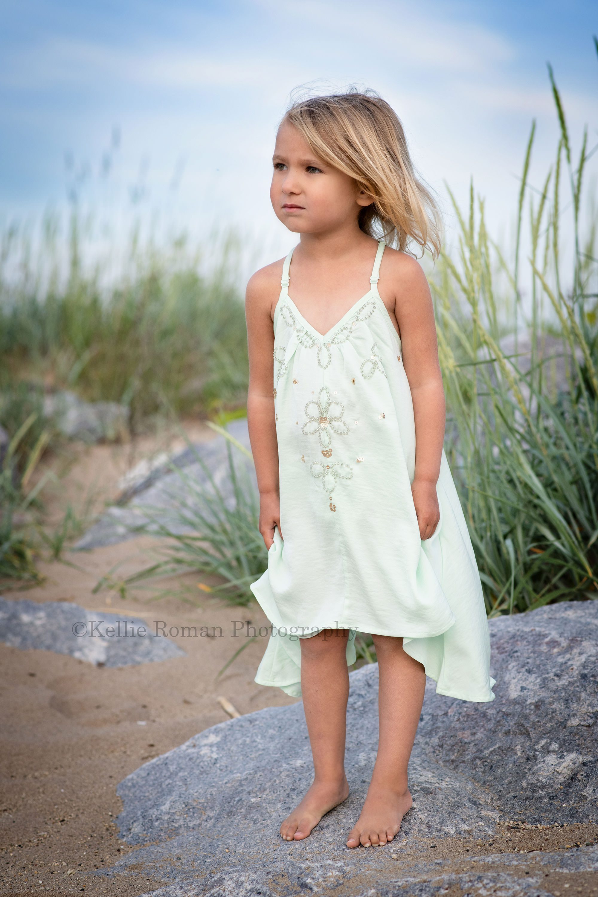 beach colors a three year old girl with blonde hair standing on a large rock in the sand on a beach she is looking off to the side and has an aqua dress on with beads she's being photographed on a milwaukee beach