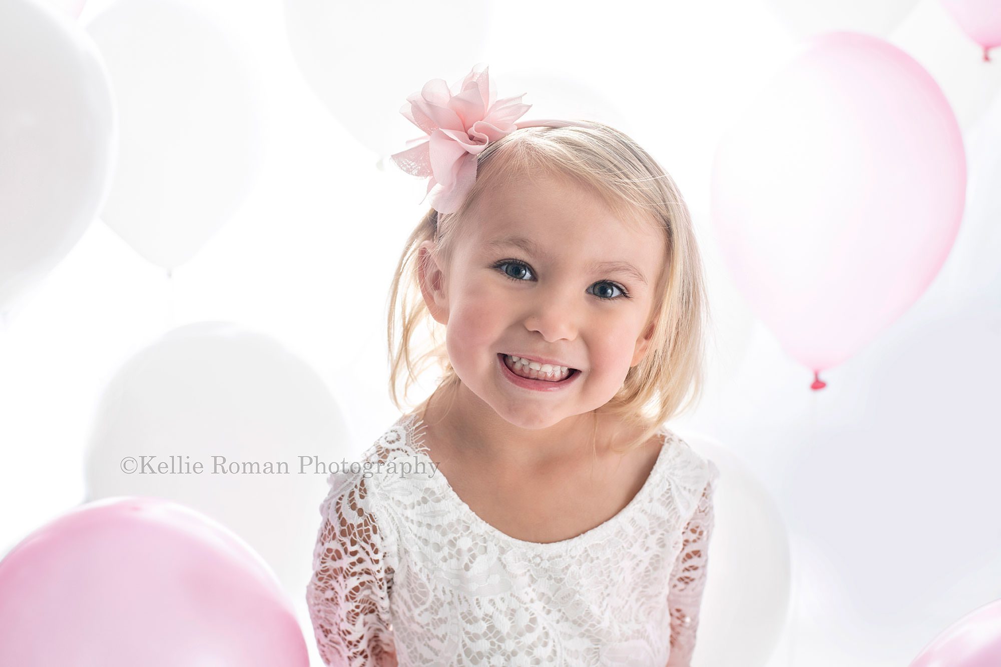 beach colors a three year old girl with blonde hair is in milwaukee photo studio she's wearing a white lace and pink tule dress surrounded by pink and white balloons she's smiling into the camera
