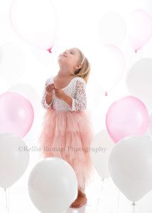 beach colors a three year old girl in a milwaukee photo studio surrounded by white and pink balloons she's wearing a frilly pink and white dress and is holding a balloon while looking up at it