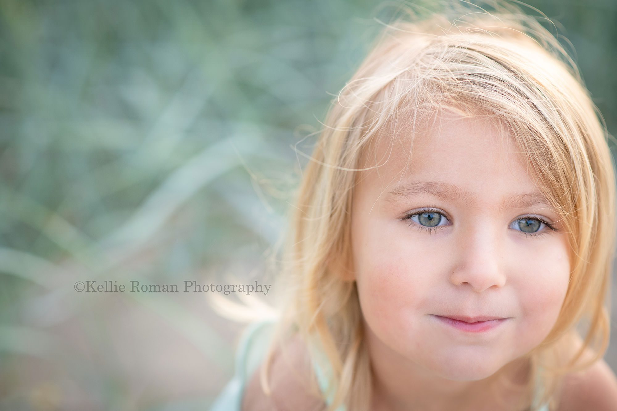 beach colors a close up photo of a three year old girl with blonde hair and green eyes she is looking off to the side and smiling with tall beach grass behind her