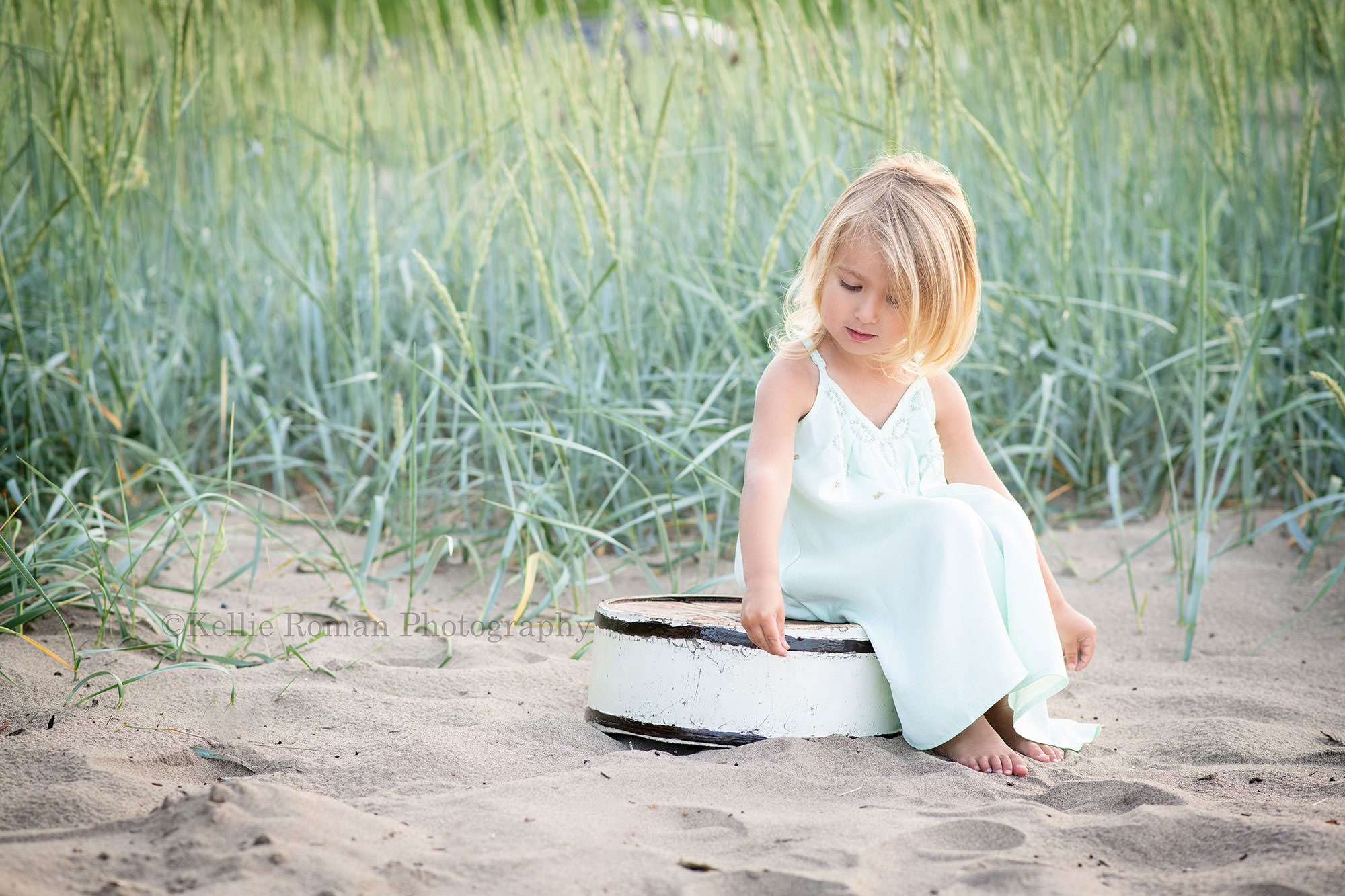 beach colors a shot of a toddler girl sitting on a white wood tub on the beach sand she has on a mint colored flowing dress and is playing with the sand while looking down there is tall teal colored beach grass behind her
