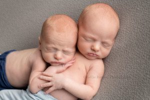 newborn twin pics two twin newborn brothers posed onto of a grey blanket in Milwaukee photography studio one boy is laying on his back and the other is on his tummy ontop of his brother they are holding onto each others arms and both are sleeping