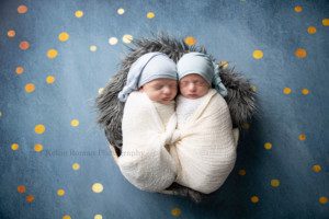 newborn twin pics shot of twin brother wrapped in ivory swaddles they swaddles are knotted together in the center they are sleeping wearing blue hats and laying in a tub with a grey fur underneath the tub is onto of a navy and gold polka dot backdrop