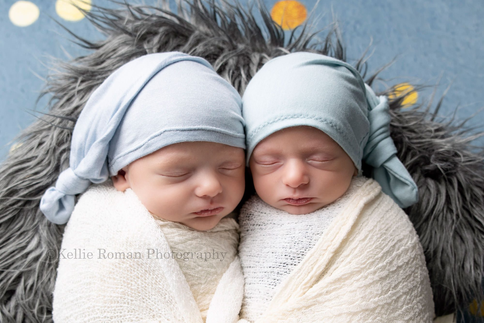 newborn twin pics two newborn twin brothers are sleeping in milwaukee photographers studio they are wrapped in ivory fabric and they both have light blue stretchy hats on they are laying on top of grey fur