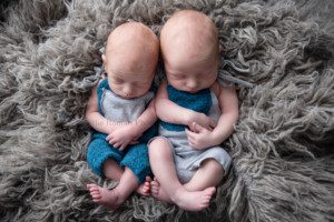 newborn twin pics newborn brothers wearing rompers that are blue and grey posed laying in a bucket with a grey fur under them they both have their legs and feet crossed and are sleeping in a milwaukee photo studio