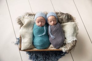 newborn twin pics in milwaukee photographer studio boys are wrapped in grey and blue swaddle wraps with blue bonnets they are both sleeping in a wood crate covered in brown and cream fur fabric the wood crate is onto of a cream wood backdrop