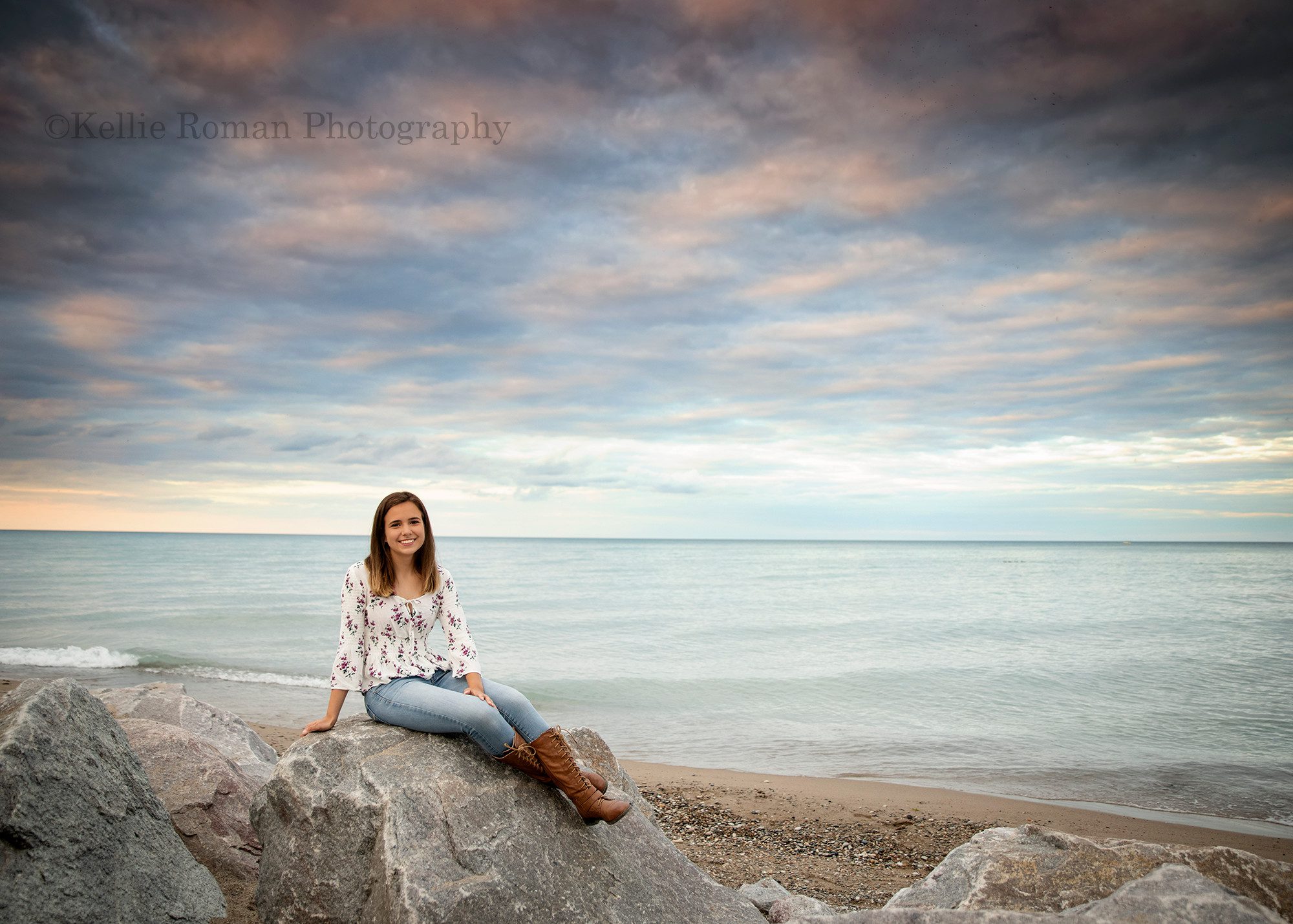 lighthouse senior pics a senior girl sitting on a massive boulder with her legs crossed there are many large rocks around her and a lighthouse behind her the clouds are very dramatic she is looking and smiling at the camera