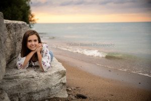 lighthouse senior pics a girl laying on her stomach on a massive rock along the shores of Lake Michigan in milwaukee Wisconsin she is smiling at the camera with her hands under her chin the lake is a shade of teal and the sun is setting