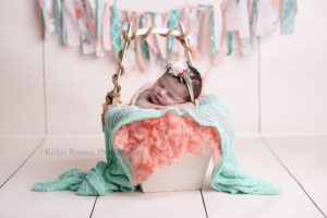 newborn pics a baby girl laying in a white bucket with a handle she is posed with her hands under her chin the bucket has layers of fabric that are teal and pink the backdrop is fake cream wood planks and there is a fabric banner with shades of pink and teal