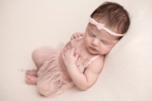 baby girl newborn laying on her back onto of a cream colored blanket she's posed with her hands on her chest and is wearing a light pink tutu romper she has a small light pink headband in her hair and is sleeping