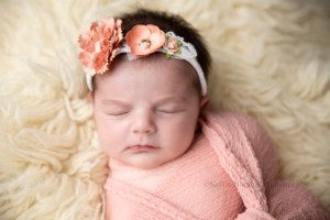 newborn pics a baby girl in a pink fabric wrap with a knot in the center is sleeping on top of cream colored fur she is wearing a floral headband in a Milwaukee photographer studio