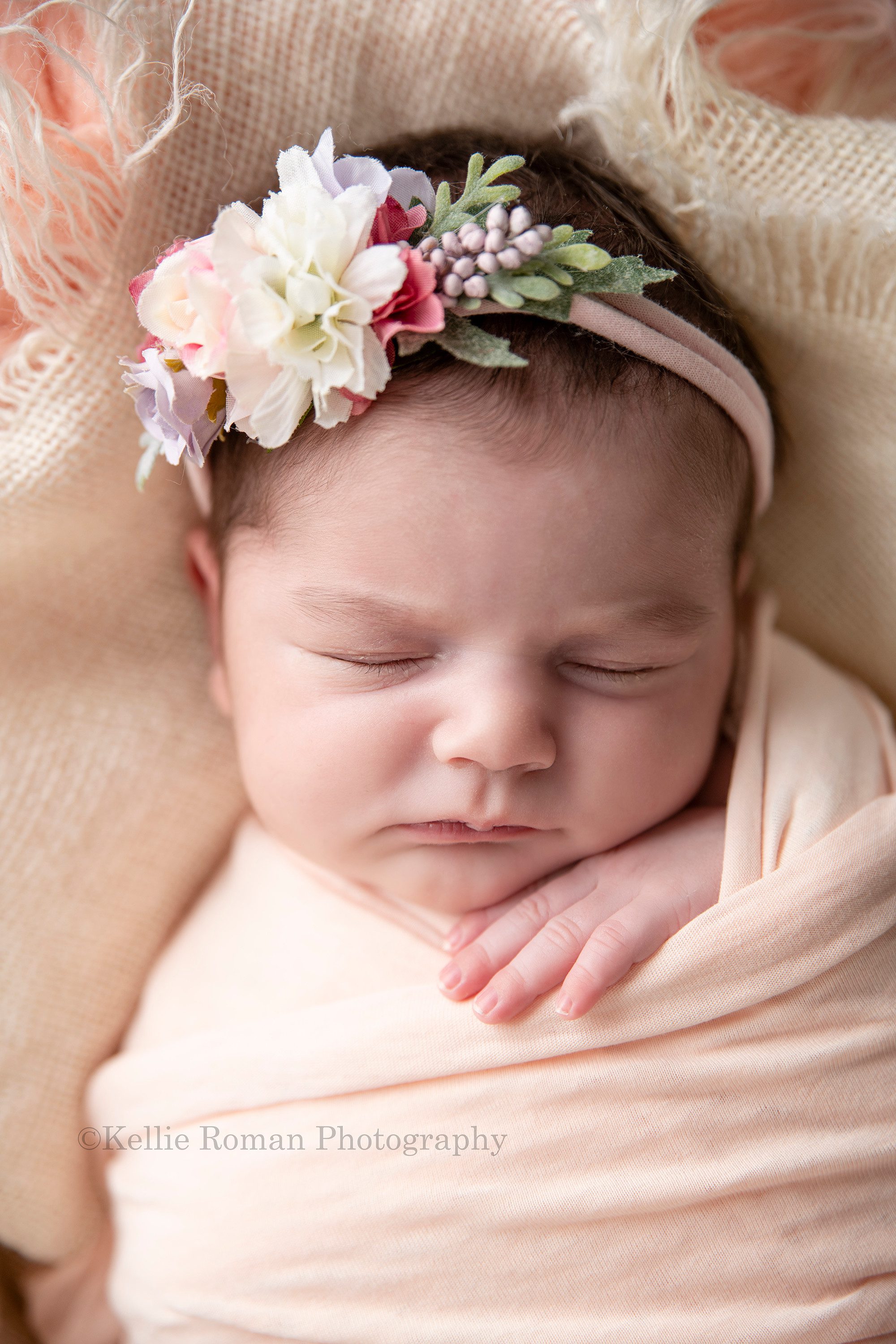 newborn pics a close up of a baby girl wrapped in a peach swaddle she is sleeping and has one hand out of the swaddle wrap she has a floral headband on