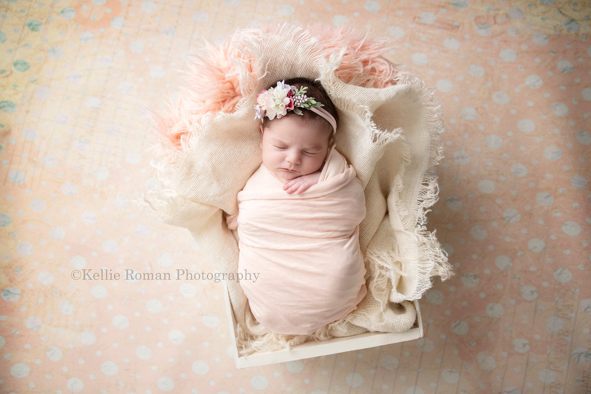 newborn pics a baby girl wrapped in a light peach fabric she is laying onto of cream burlap in a white wood crate the crate is on top of a polka dot peach backdrop the baby has very dark hair and is wearing a headband