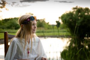 boho inspired a young blonde girl sitting on a chair looking off to the side she is wearing a white lace top with a gold necklace and flower headband