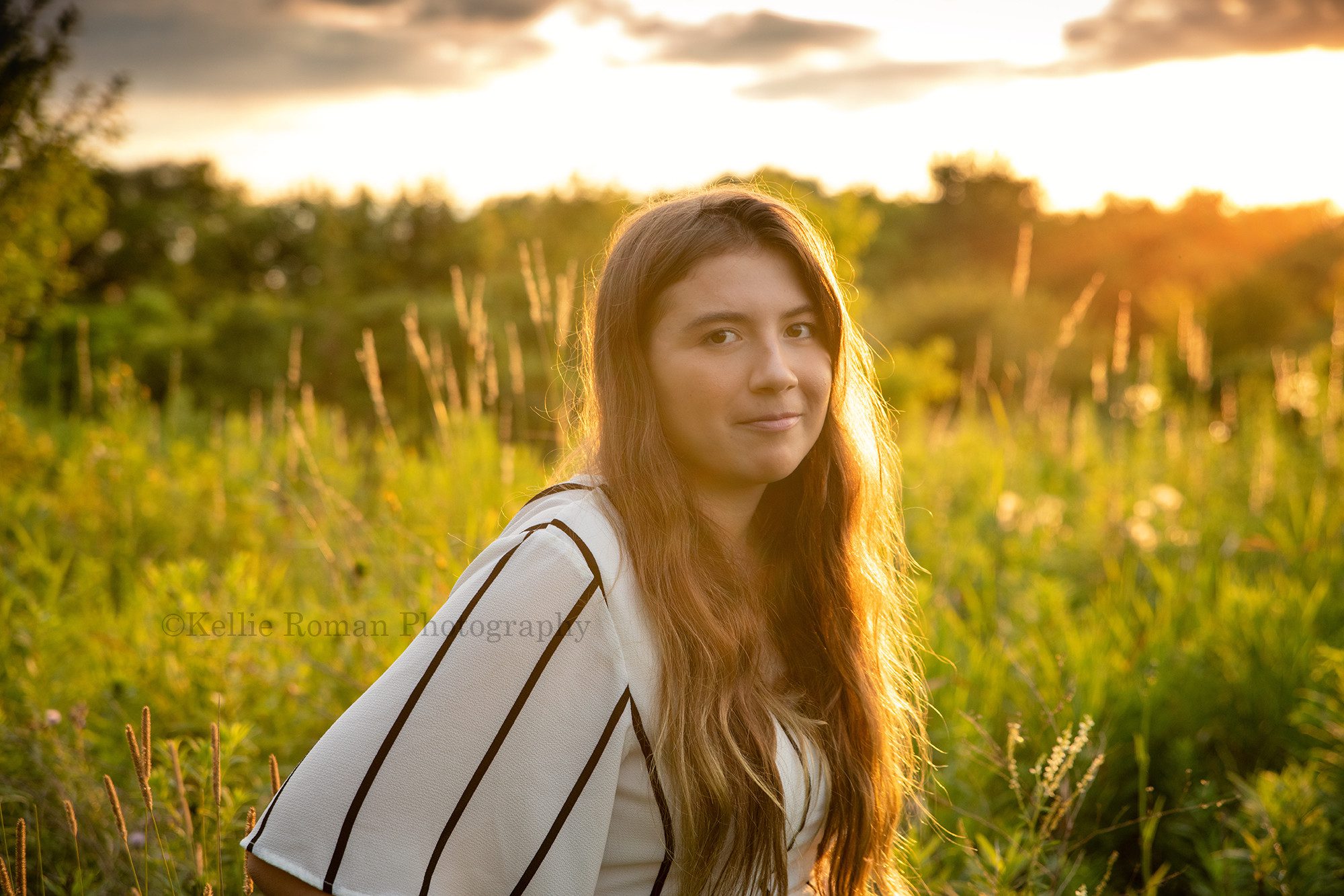 boho inspired a teenage girl looking into the camera she is standing in a field of tall grass with the setting sun behind her she has on a white and black outfit with stripes