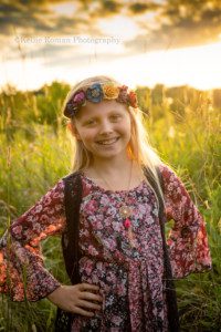 boho inspired a young girl looking at the camera standing in a field of tall green grass she is wearing a patterned maroon dress and a flower felt headband the sun is making her blonde hair glow