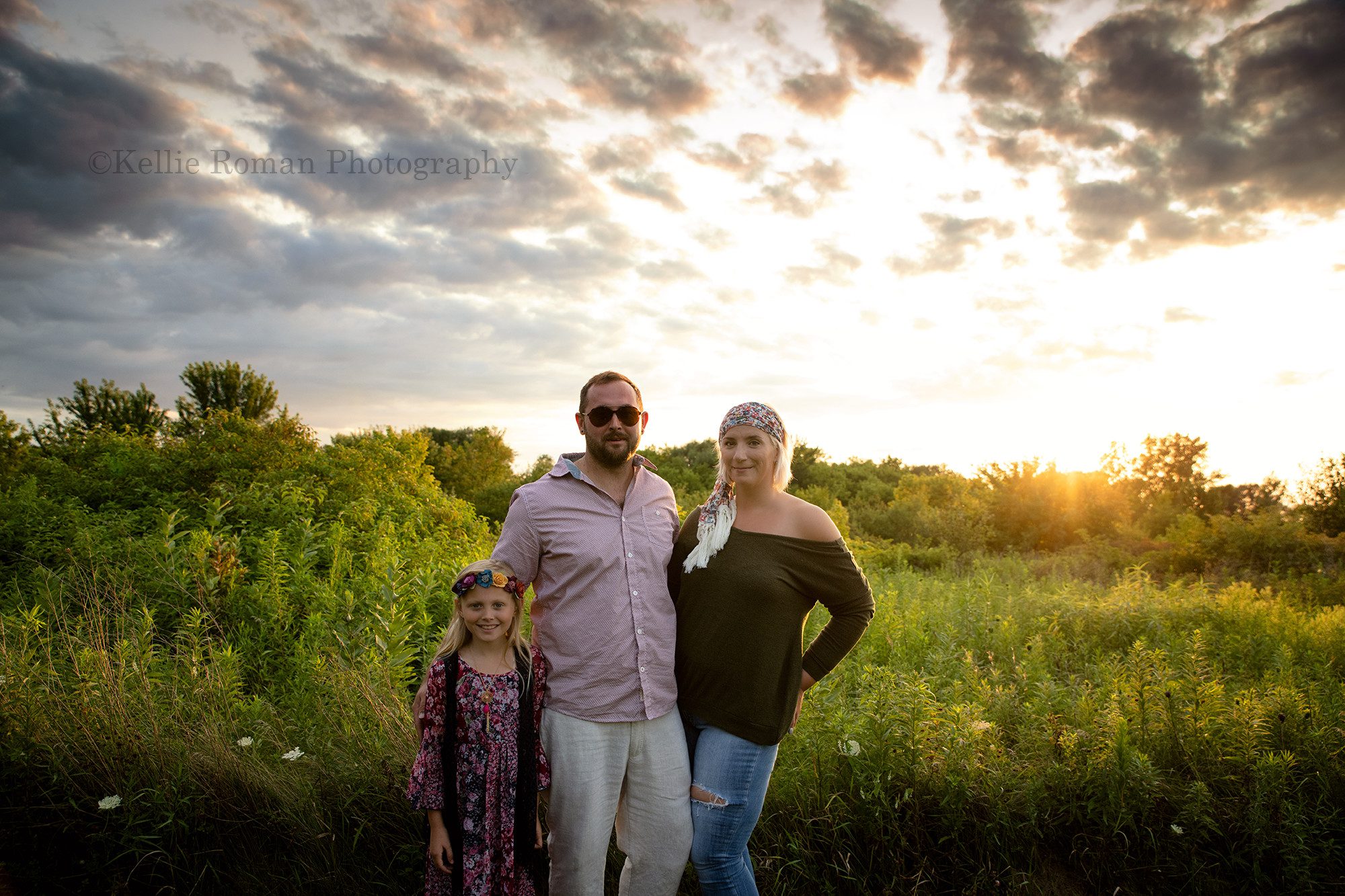 boho inspired family session with three family members standing in a field of tall grass and flowers in Wisconsin they are wearing dark colors and flower headbands the sun is setting behind them