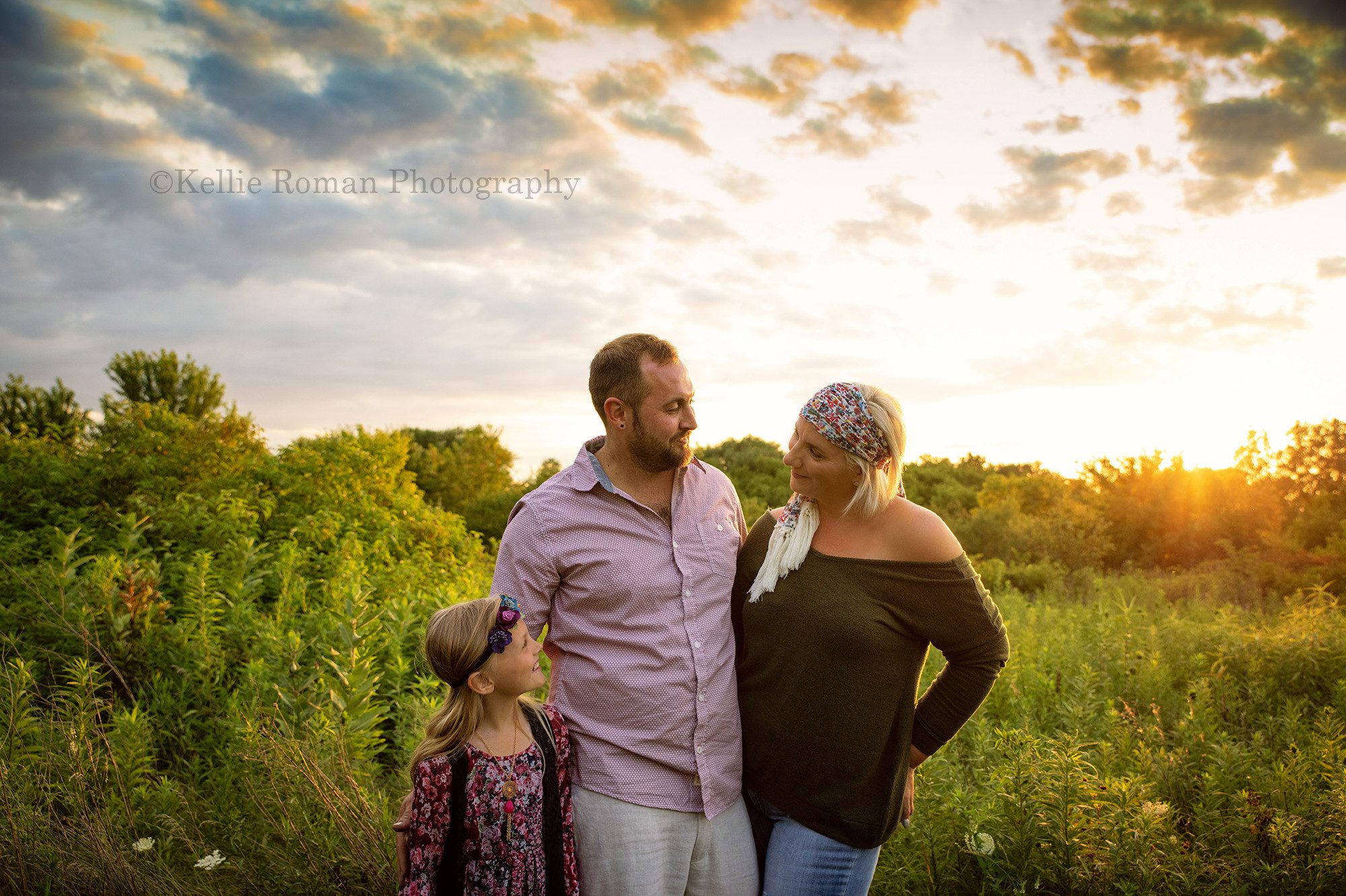 boho inspired family session a family of three standing in a field of tall green grass and flowers with the sun setting behind them they are wearing shades of maroon and dark green they are looking at each other and smiling