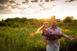 boho inspired a father spinning his young daughter around in his arms in a field of tall grass and flowers the sun is setting behind them and glowing yellow the girls blonde hair is flying off to the side and they are both smiling and laughing