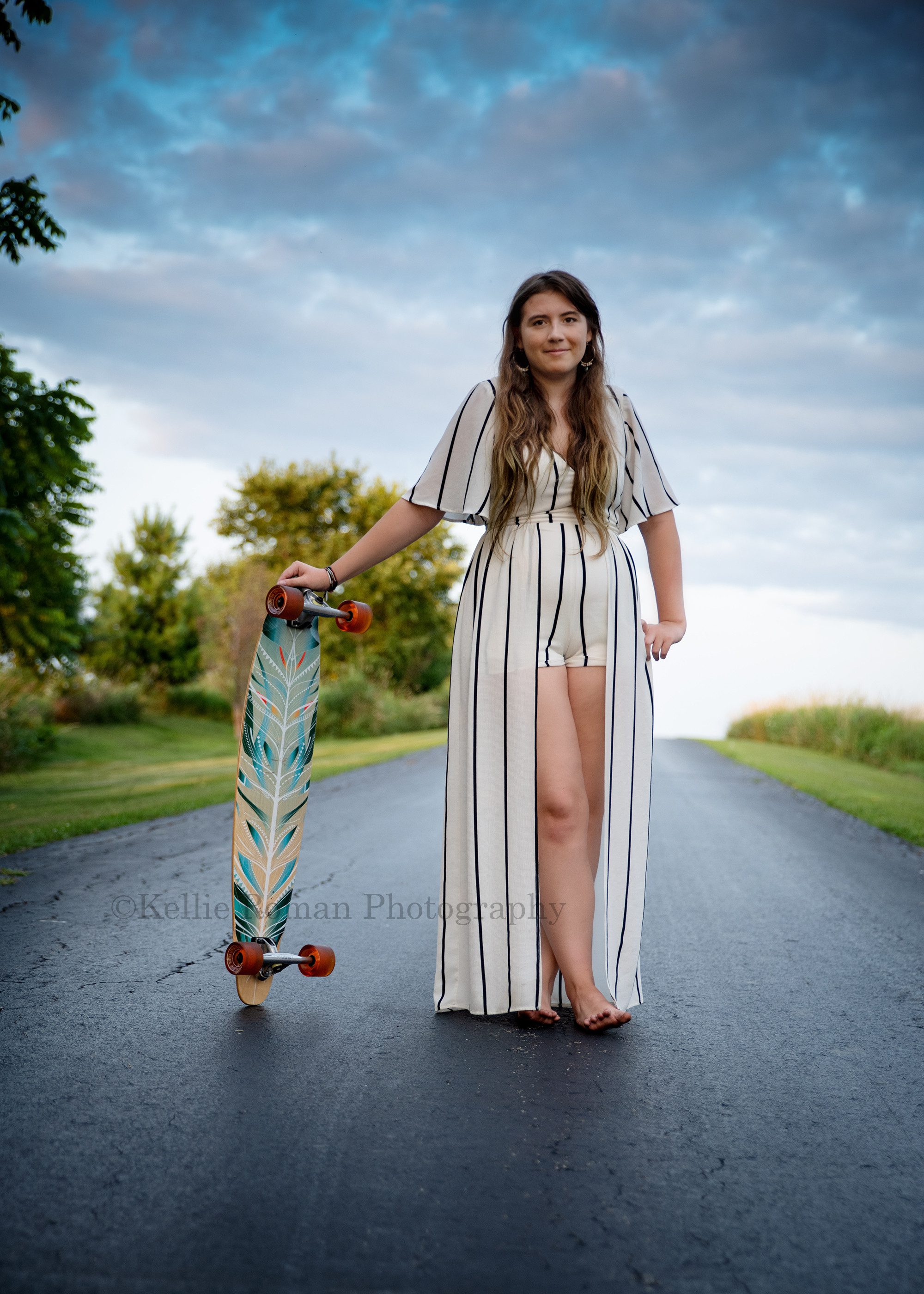 boho inspired a teenage girl with very long hair is stand on a blacktop driving she has her feet crossed in front of her and is leaning to one side on a very colorful skateboard that is standing upright she has a romper shorts outfit on