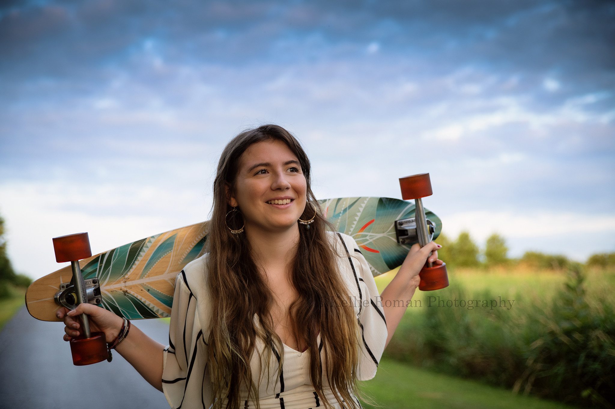 boho inspired a teenage girl with very long hair standing on a black driveway she is looking up and off to the side smiling and is holding a colorful skateboard behind her shoulders she has on a white long romper with black stripes