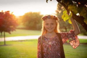 boho inspired a photo of a young girl with the sun setting behind her looking into the camera she has a maroon dress on with a gold necklace and vest she is holding onto a tree brand and wearing a floral headband