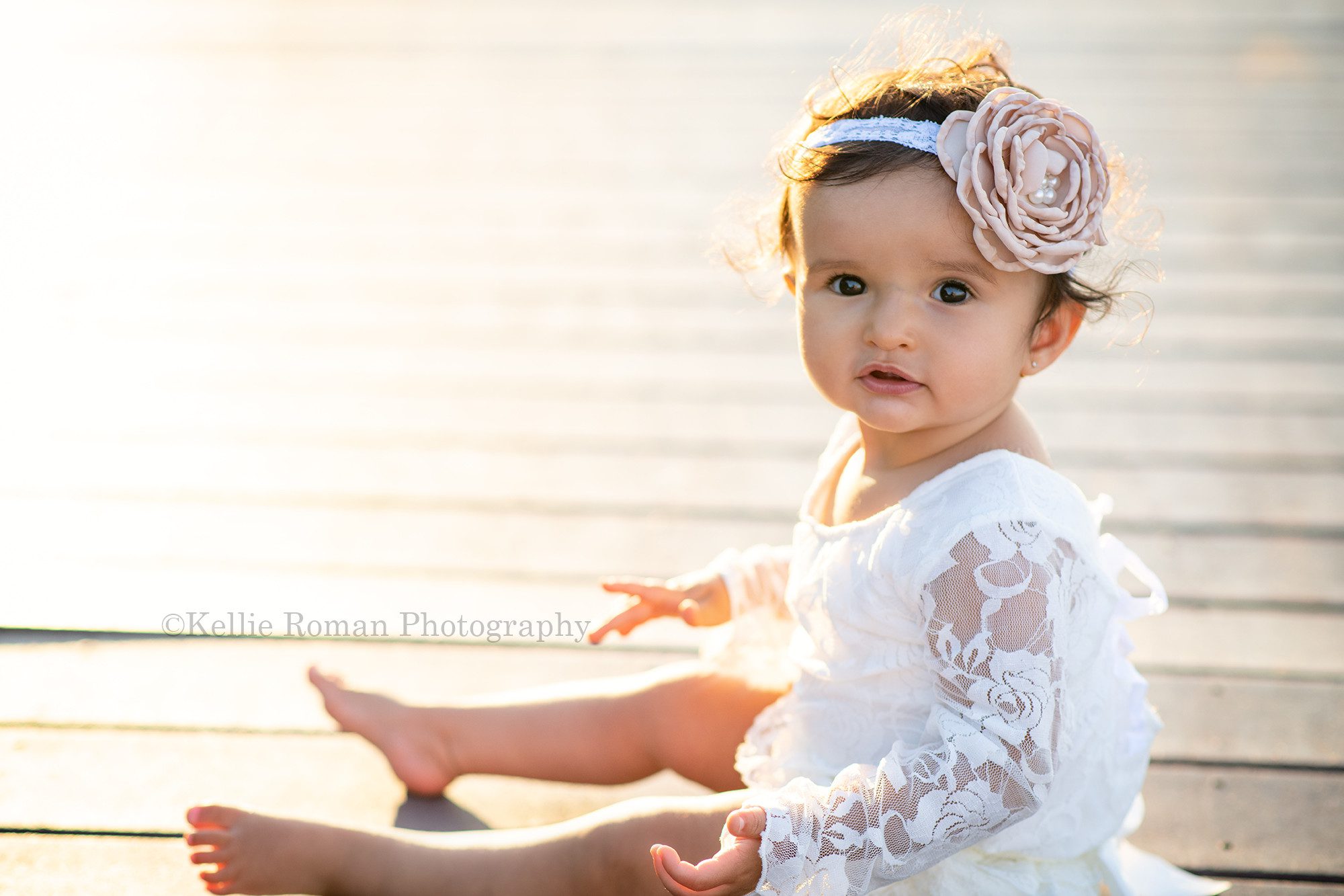 beach cake smash a one year old girl sitting on a wood walk way with the sun shining behind her she is looking into the lens of the camera and is wearing a lace ivory romper with a pink headband