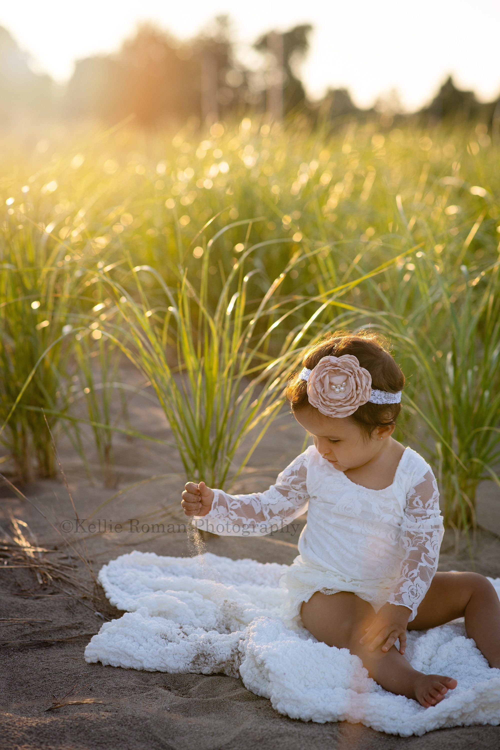 beach cake smash a one year old girl sitting on top of a white blanket wearing a lacy white romper she is playing with the sand and it's falling out of her hand while the sunshine is shining though it she has tall green beach grass behind her