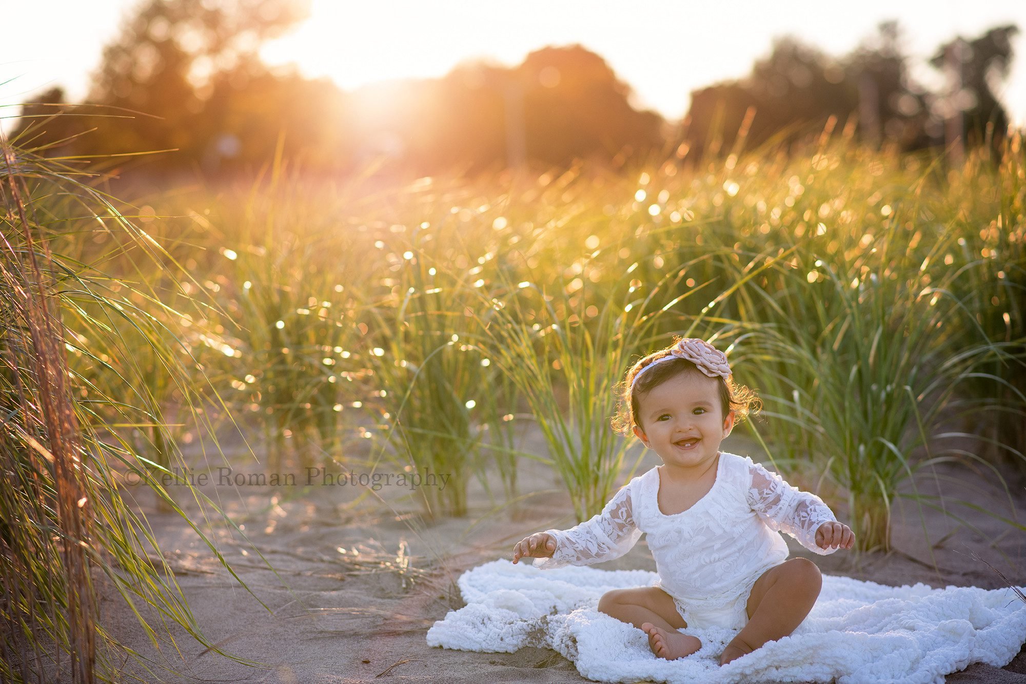 beach cake smash a little girl wearing an ivory romper made of lace is sitting on top of a white blanket on a beach the sunshine is shining through tall beach grass behind her