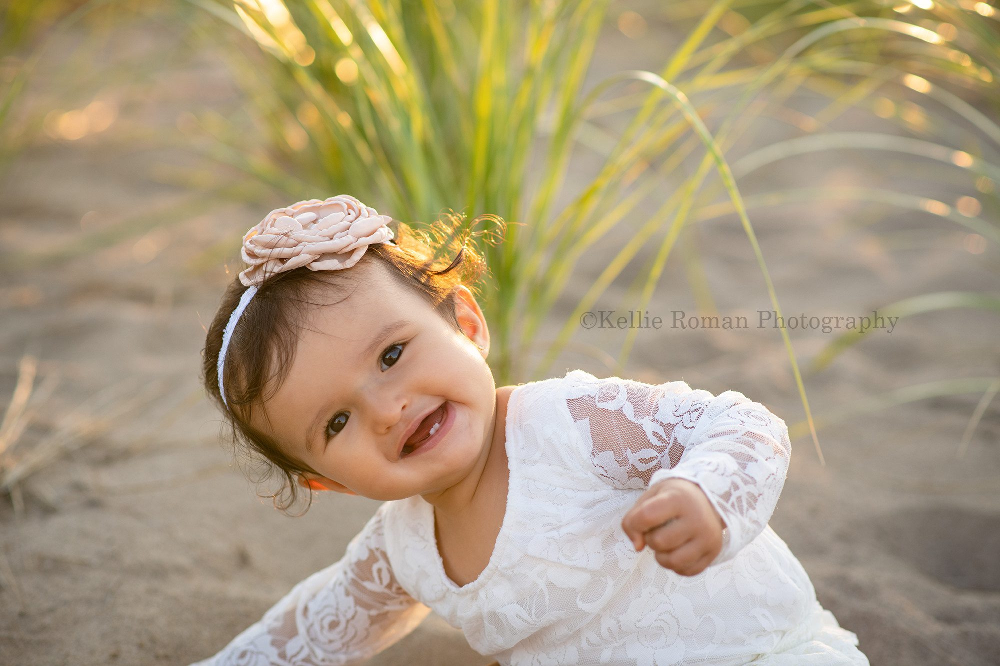 beach cake smash a little girl wearing a lace ivory romper is sitting on the beach in front of tall green beach grass she is leaning over and smiling into the camera the sun is shining from behind her