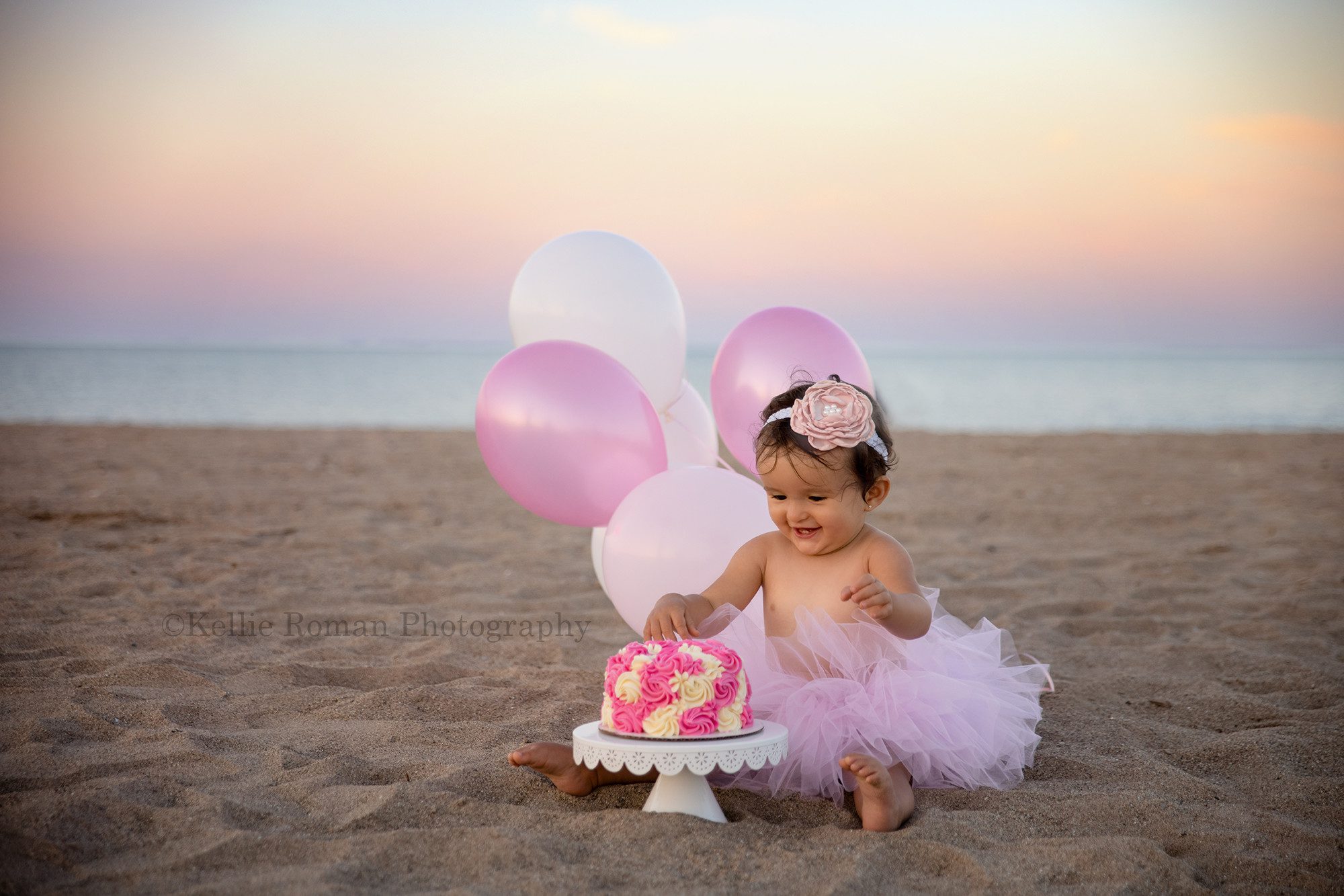 beach cake smash a little girl who's celebrating her first birthday is on a beach in kenosha she has a pink tutu on and is ready to smash her hands into a pink and white frosted cake she has balloons behind her