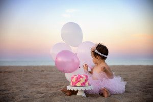 beach cake smash a one year old girl sitting on a beach looking at pink and white balloons that are behind her she has a pink and white cake in front of her on a white cake stand