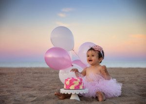 beach cake smash a one year old girl smiling really big showing her two bottom teeth she is sitting on a sandy beach in Wisconsin and is wearing a pink tutu with a white and pink frosted cake in front of her she has a headband on and pink and white balloons behind her