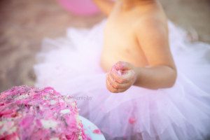beach cake smash a close up photo of a one year old girls hand with frosting on it she is on a beach in kenosha Wisconsin and is wearing a pink tutu sitting behind a pink and white frosted cake