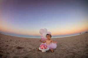 beach cake smash a one year old little girl sitting on a sandy beach in kenosha Wisconsin wearing pink tutu behind a pink and white cake she has pink balloons behind her and frosting is on her hands the sky is shades of pastel above the water