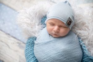 baby pics a newborn baby boy in a blue swaddle wrap is sleeping in a wood crate there are layers of cream fur and blue knit blankets in the bucket and the baby has a blue hat on with a button on it