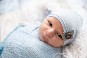baby pics a newborn boy wrapped in a light blue swaddle wrap with a light blue hat on is laying on his back in milwaukee photographer studio he has is eyes open and is looking to the side