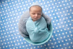 baby pics a newborn baby boy is wrapped in a blue swaddle and laying upright in a blue bucket with a grey fabric in it he has a bonnet on and the bucket is onto of blue polka dot backdrop he's being photographed by a milwaukee photographer