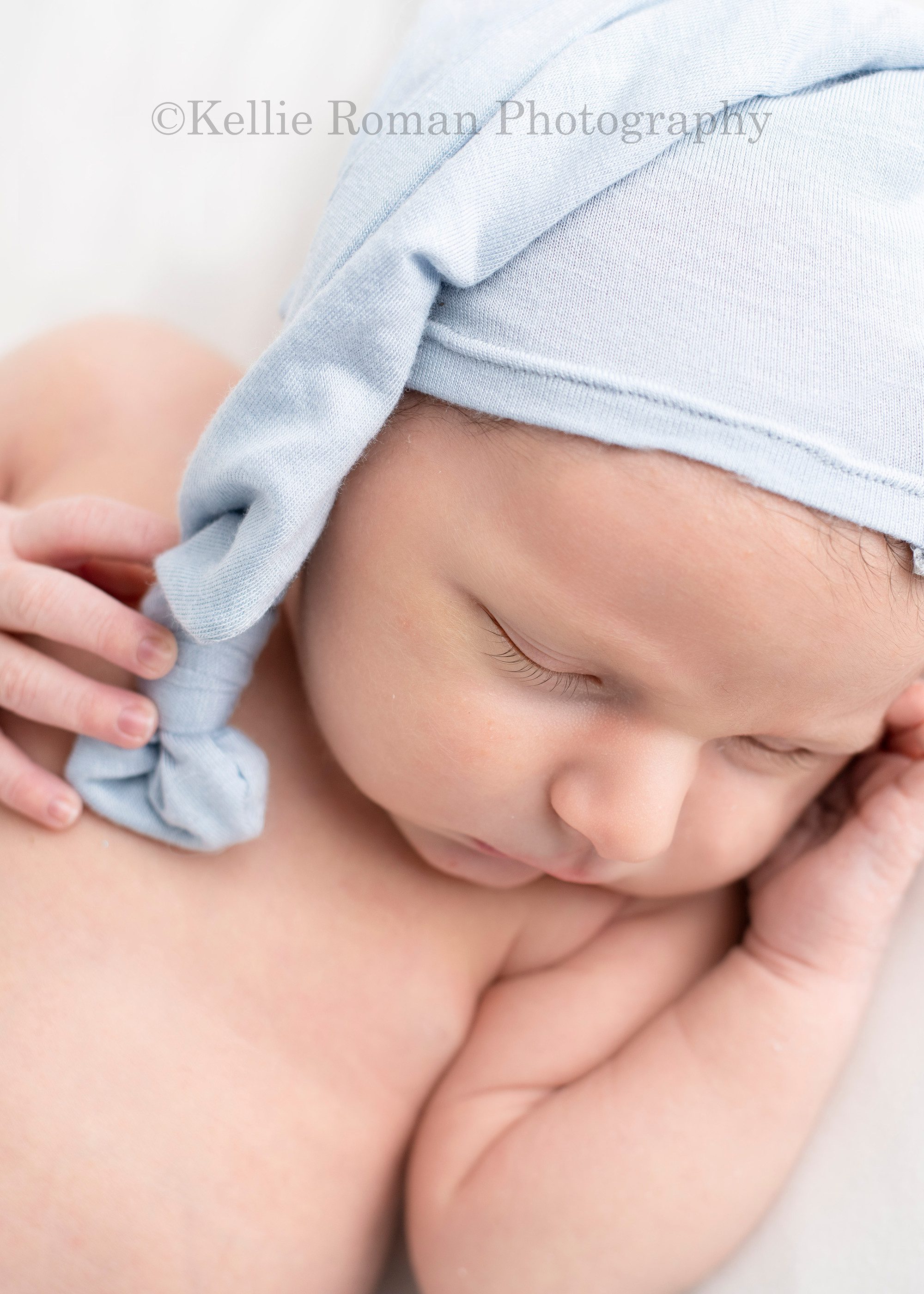 baby pics a newborn boy laying on a cream colored blanket in a milwaukee photography studio he is asleep on his back and has a light blue hat on with a knot at the top of it the shot is a close up of his face and hand