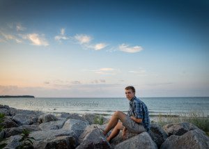 oak creek senior guy sitting on large rocks at Lake Michigan shore in oak creek he is wearing cargo shorts and a blue plaid shirt the sky is shades of pastel behind him