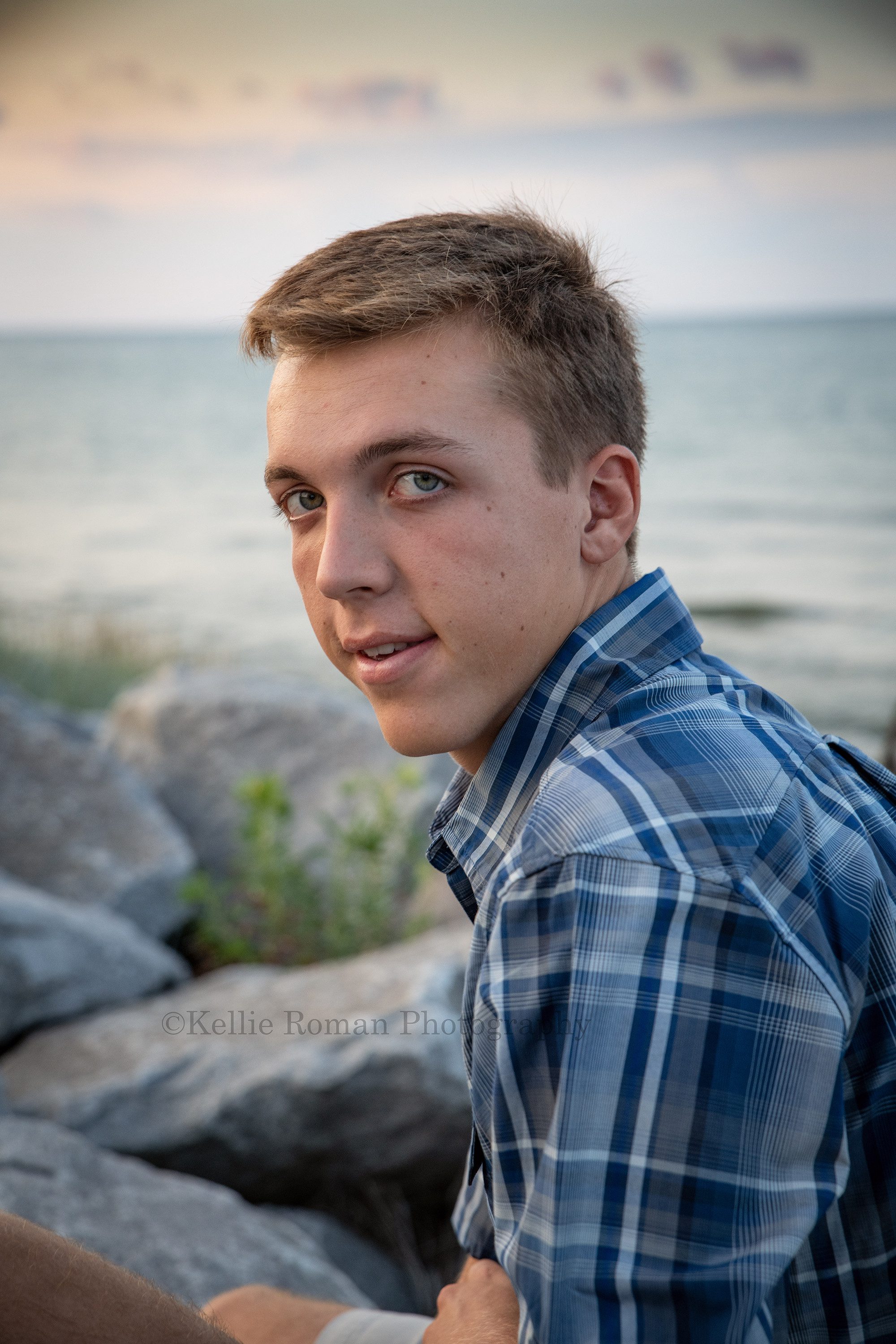 oak creek senior student in milwaukee county park sitting on large rocks in front of Lake Michigan he is looking at the camera while wearing a blue plaid shirt the sky is pastel behind him