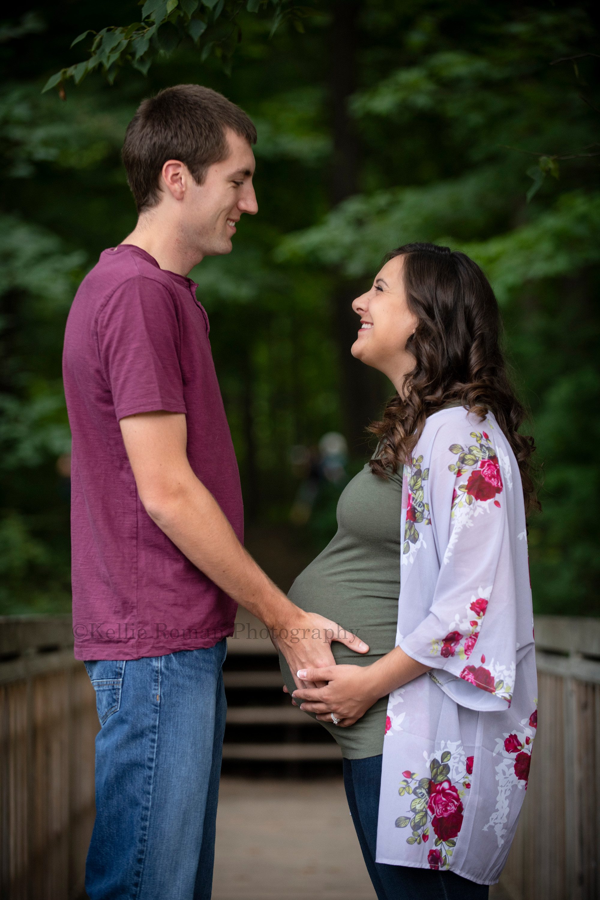 maternity gowns a husband and wife expecting their first baby standing on a wood bridge in Kenosha park they are facing each other and smiling while they have their hands on the women baby bump