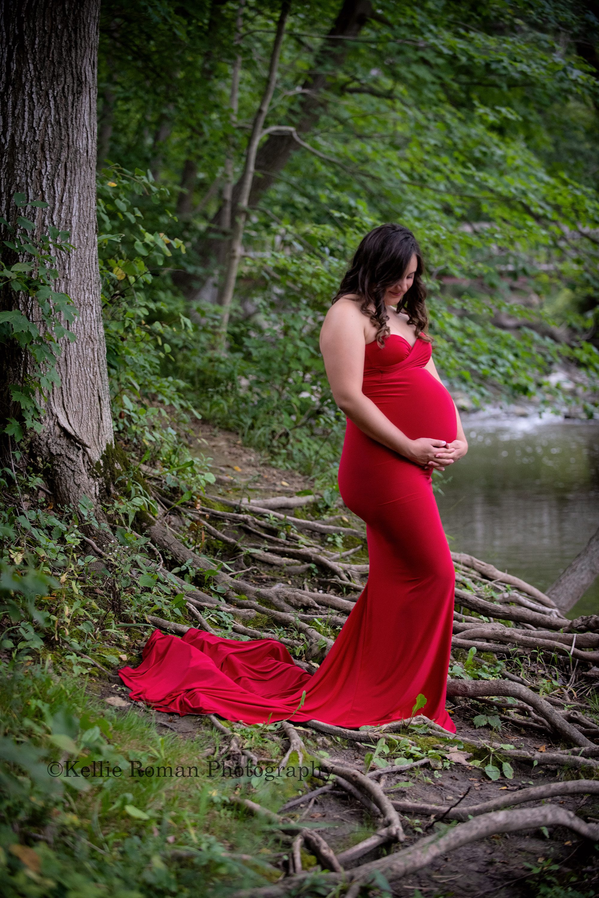 maternity gowns a pregnant women standing in a kenosha park on top of rugged tree roots near a river she's wearing a red tight maternity gown with a train and is looking down at her belly