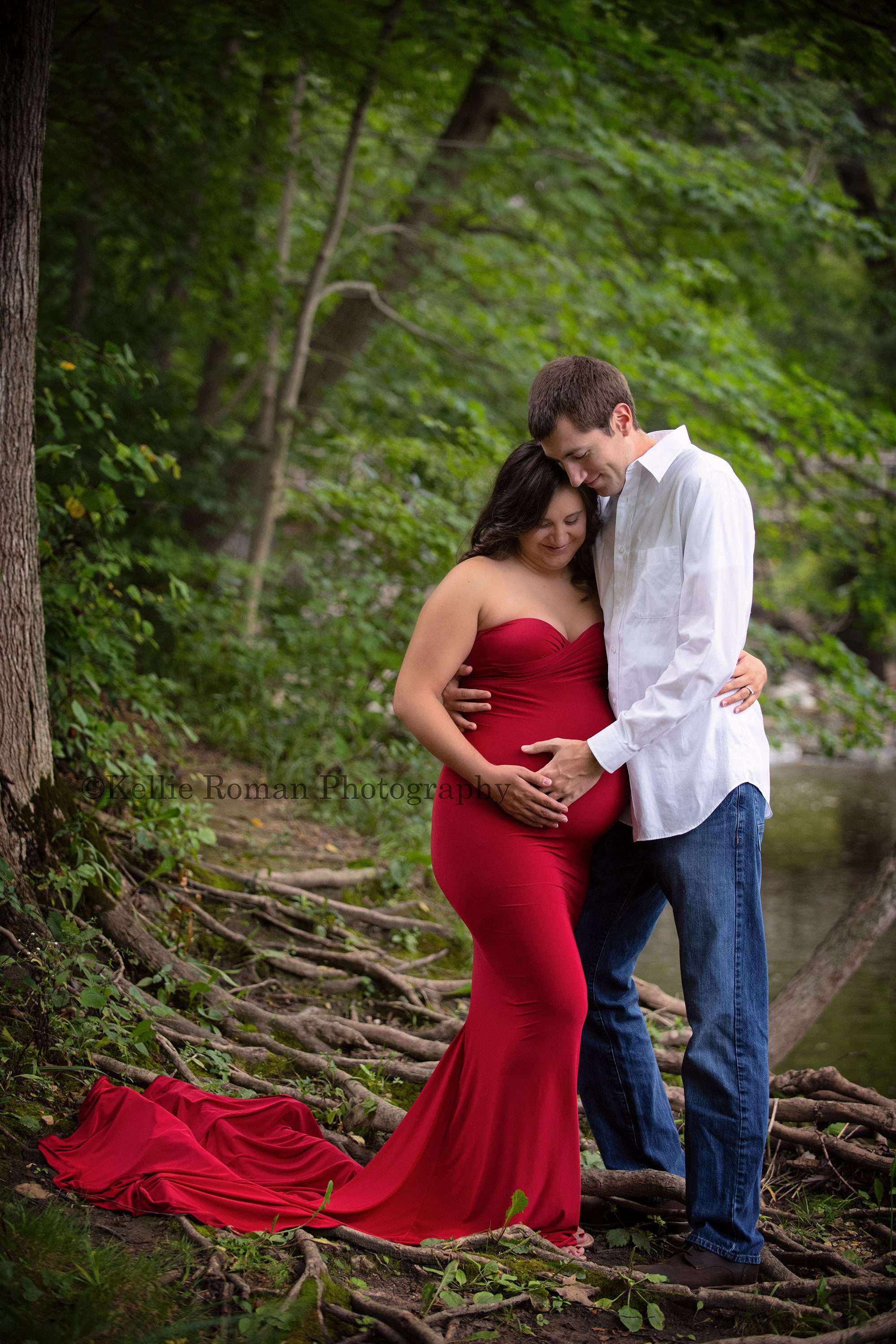maternity gowns a couple expecting their first child in kenosha standing in a park near a river they are standing on top of wild tree roots they the women is in a tight fitted red maternity gown with a train draped over the roots the man is wearing jeans and a white shirt they have their hands on women belly and are looking down smiling