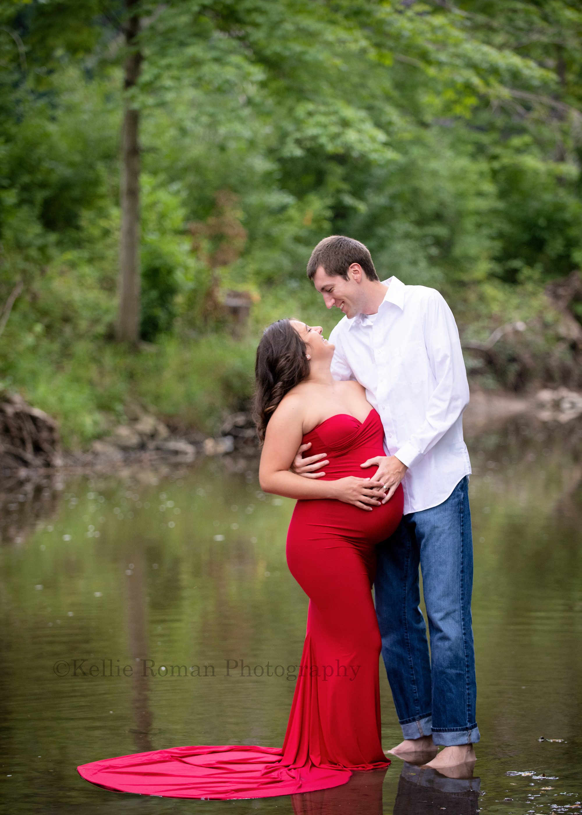 maternity gowns a husband and wife standing in a river in kenosha Wisconsin the women is pregnant and wearing a tight red gown her husband has on jeans and a white shirt they are looking at each other and smiling