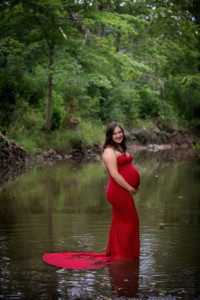 maternity gowns a women wearing a tight red maternity gown with a train is standing in a river in kenosha Wisconsin she is looking at the camera smiling and holding the bottom of her belly