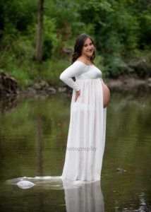 maternity gowns a women expecting her first child is standing in a river in kenosha Wisconsin with a white sheer gown on the gown has a slit up the front and her bare belly is showing through she has her hand on her hip and is looking at the camera