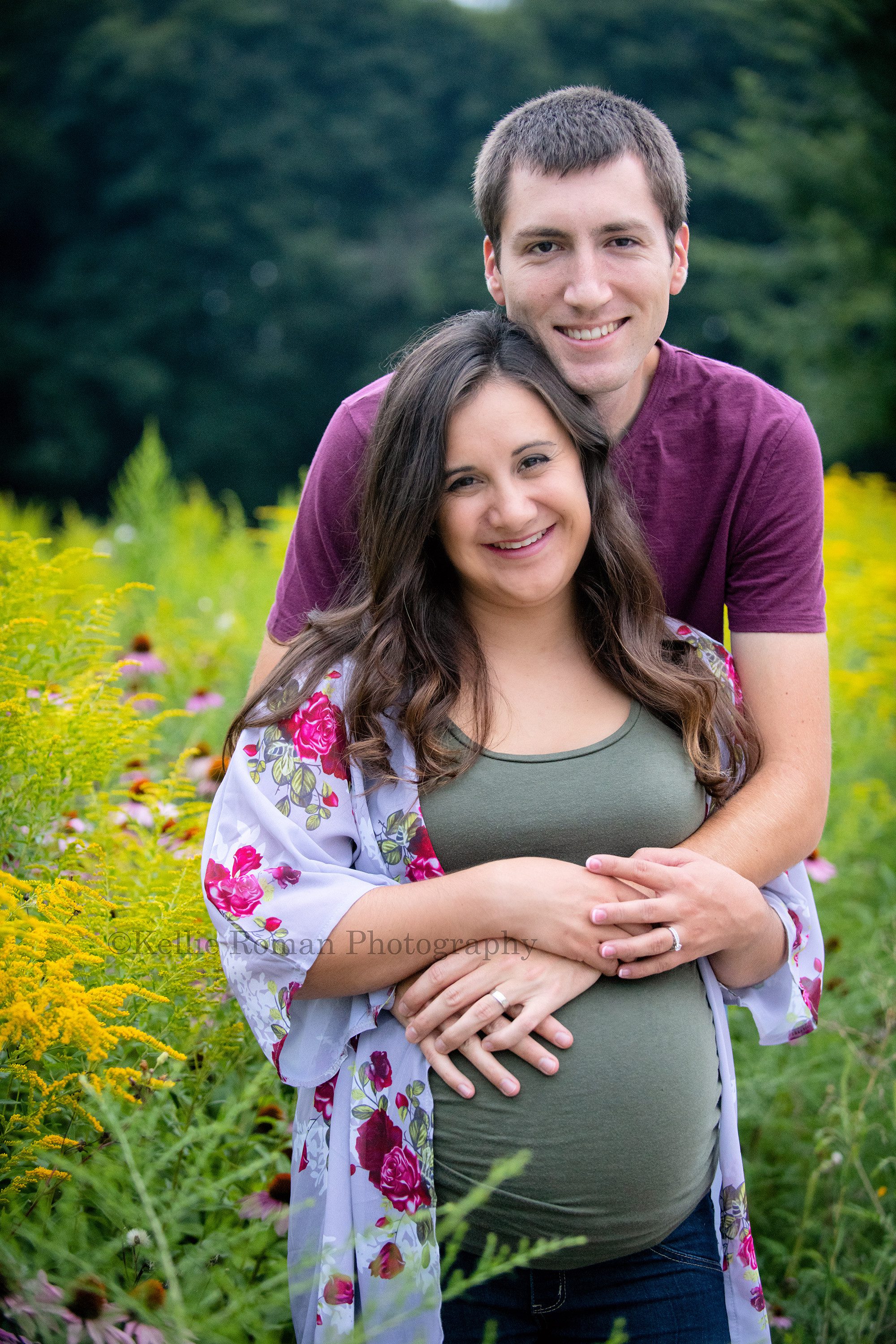 maternity gowns a couple in kenosha Wisconsin expecting their first child are standing in a field of yellow pink and green flowers they are looking at the camera smiling and they are wearing shades of green white and maroon with jeans