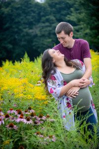 maternity gowns a couple standing in a field of flowers and tall grass looking at each other the women is very pregnant and they are wearing shades of green and maroon