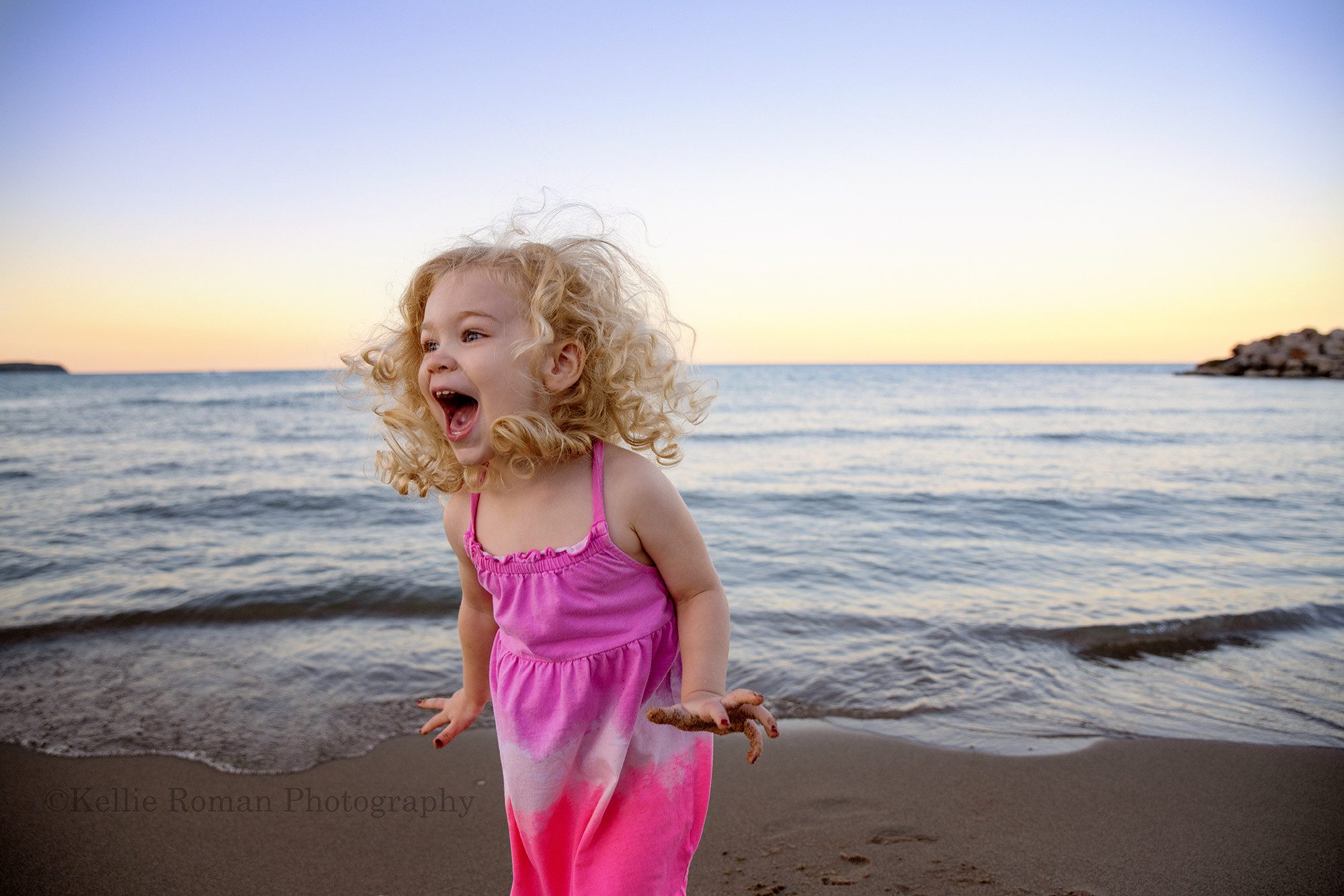 bubbles at the beach a three year old girl on a beach in milwaukee is jumping in excitement she has on a pink dress and has long curly blonde hair which is in front of her eyes the sky and lake water are shades of pastel with the sun setting
